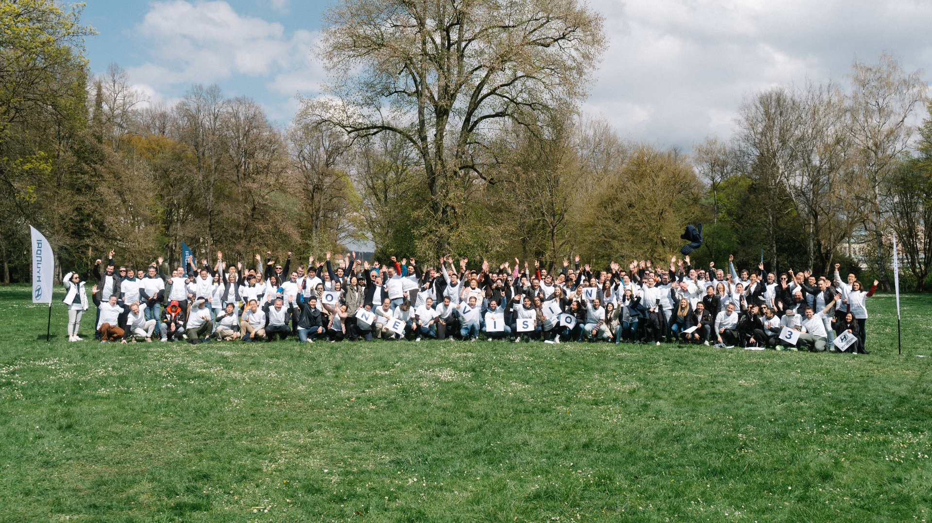 A large group of enthusiastic people gathered outdoors for an event photography shoot on a sunny day, with some individuals holding banners or signs, celebrating a shared event.