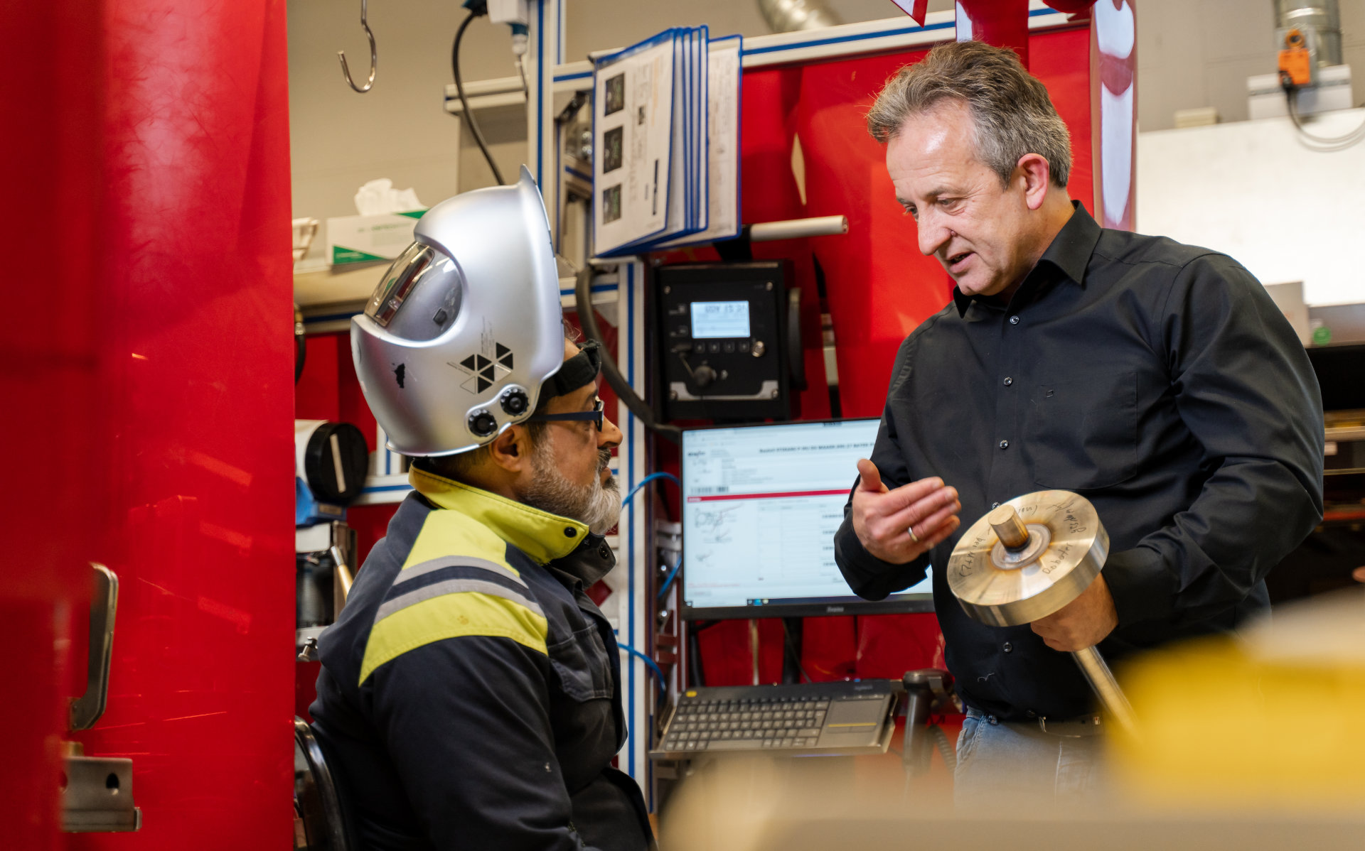 A worker with a helmet and visor engaged in a discussion with a supervisor holding a metal part in an industrial setting with machinery and computer equipment in the background.