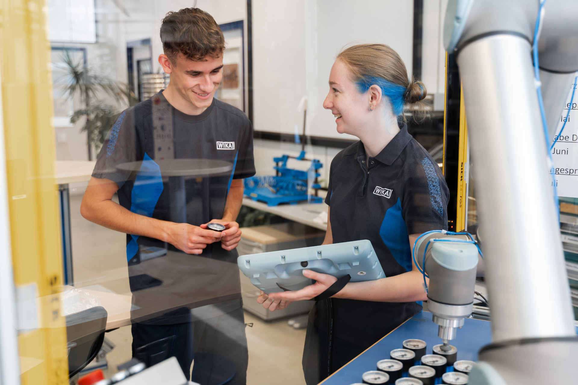 Two technicians in matching uniforms smiling and inspecting a part next to a robotic arm in a modern industrial environment.